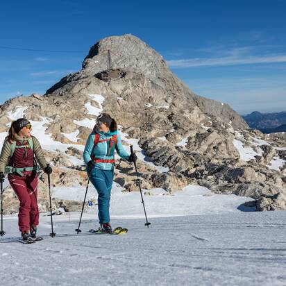 Zwei Frauen gehen mit Skitouren-Ausrüstung vor felsigem Gipfel bei Sonnenschein | © H. Steiner
