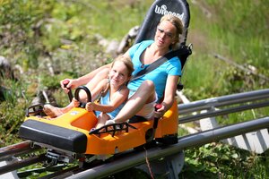 Mother and daughter ride the Rittisberg alpine coaster together, smiling while racing downhill in a summer toboggan car. | © photo-austria