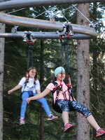 Two children glide safely in harnesses on the Flyline ride through the forest at Rittisberg, surrounded by tall pine trees. | © Erlebnis Rittisberg