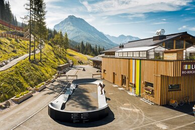 Skater auf einem Pumptrack vor dem Holzgebäude des Bio-Bergrestaurants in alpiner Landschaft. | © Shuttleberg