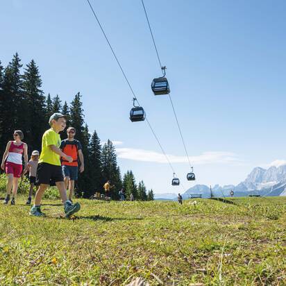 Eine Familie wandert bei Sonnenschein auf einer Almwiese unter schwebenden Gondeln mit Blick auf die Berge von Ski amadé.