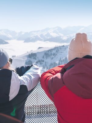 Zwei Personen in Winterkleidung stehen auf Terrasse und schauen auf verschneite Berge mit Skiern im Vordergrund