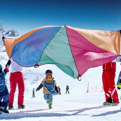 Kinder in Best Learn2Ski-Westen spielen unter buntem Schwungtuch auf verschneiter Piste mit Skilehrern im Hintergrund