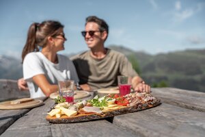 Couple enjoys alpine snack with cheese, bacon, juice on wooden table and mountain view | © Skigastein, seven frames