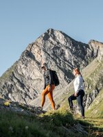 Two women hiking uphill at Graukogel, with a rugged rocky mountain peak in the background under clear blue sky | © Skigastein, seven frames