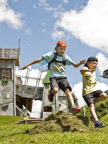 Two kids jump over a grassy mound in front of a fantasy play tower at Geisterberg adventure park in St. Johann, Salzburg | © sanktjohann.com | Mirja Geh