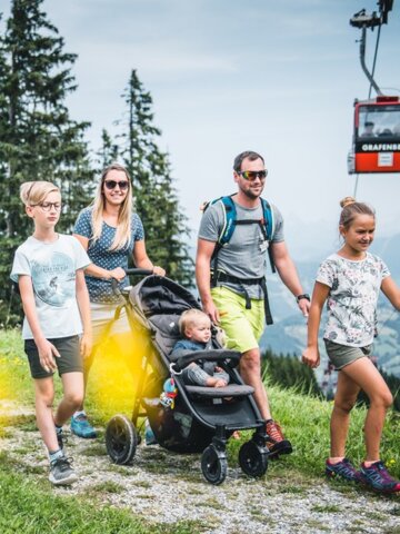 Family with stroller hiking along Grafenberg trail, a red gondola passes by in the background of the scenic view | © Snow Space Salzburg