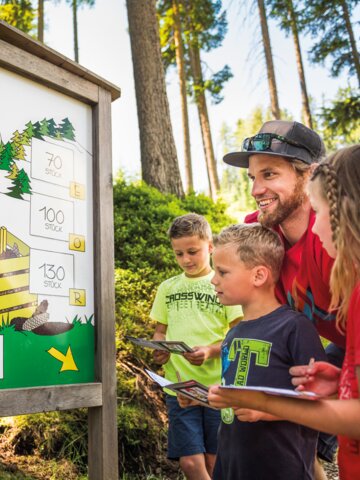 Family with three kids stands at forest quiz board with cones, all holding booklets, smiling and engaged in guessing