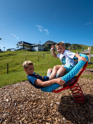 Two laughing kids swing on rope seat near Gamskogelbahn Zauchensee during summer vacation. | © Zauchensee Liftgesellschaft