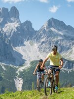 Two cyclists ride across a green meadow in Filzmoos with steep grey mountains under a blue sky | © Lorenz Masser