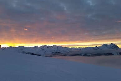 Verschneite Berghänge mit orangefarbenem Sonnenaufgang und Wolken über Mittelstation