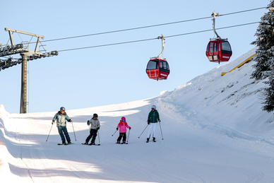 Vier Skifahrer, darunter zwei Kinder in bunter Kleidung, fahren auf präparierter Piste, rote Gondeln im Hintergrund. | © Gerhard Wolkersdorfer
