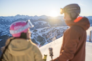 Zwei Personen in Skibekleidung mit Helm und Skibrille schauen beim Sonnenuntergang über verschneite Berge in Dorfgastein | © Dorfgasteiner Bergbahnen