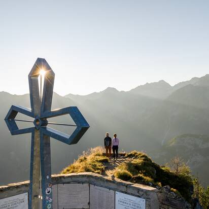 Zwei Frauen stehen am Gipfelkreuz des Graukogels, Sonnenaufgang hinter den Bergen, Blick auf alpines Panorama | © Skigastein, seven frames
