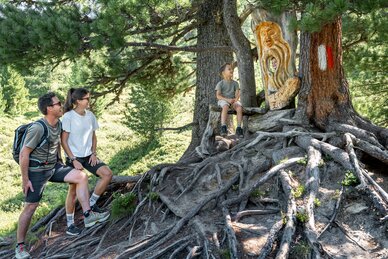 Familie am Graukogel beim geschnitzten Zirbengeist auf Wurzelbank im Wald | © Skigastein, seven frames