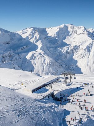 Bergstation einer Kabinenbahn von oben und daneben stehen Skifahrer und rundherum sind schneebedeckte Berge