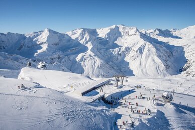 Bergstation einer Kabinenbahn von oben und daneben stehen Skifahrer und rundherum sind schneebedeckte Berge