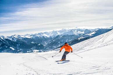 Skifahrer mit oranger Skijacke fährt eine präparierte Piste hinunter und in der Ferne sind schneebedeckte Berge zu sehen | © Gasteinertal Tourismus GmbH, Creatina