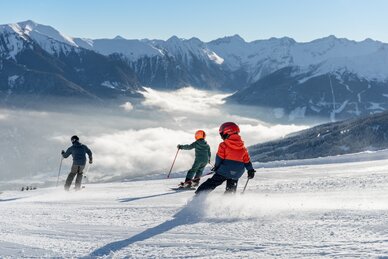 Drei Skifahrer, darunter zwei Kinder, fahren bei Sonne auf Piste mit Alpenblick | © Simon Hutter