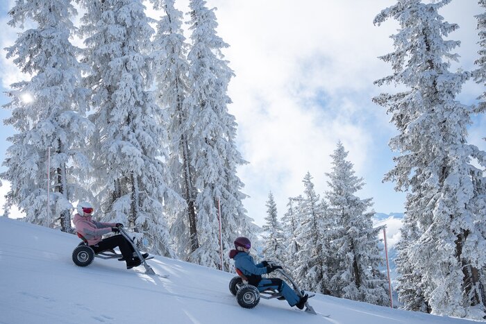Zwei Personen fahren mit Snowbikes eine verschneite Piste durch winterlichen Nadelwald hinab