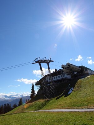 Cable car ascends to mountain station on sunny day, with green meadow, snowy mountain backdrop and clear blue sky. | © Großarler Bergbahnen