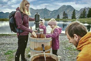 Familie mit Kindern pumpt Wasser aus Holzfässern am See in Großarl, dahinter Berge, Wolken und spiegelndes Wasser. | © Großarler Bergbahnen