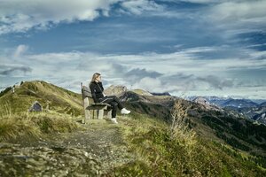 Frau sitzt auf einer Bank am Bergweg in Großarl und blickt über eine herbstliche Hügellandschaft mit dramatischer Wolkenkulisse. | © Großarler Bergbahnen