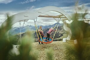 Zwei Kinder spielen auf einer großen Nestschaukel vor Bergkulisse in Großarl, Holzhackschnitzel am Boden, Wolken am Himmel | © Großarler Bergbahnen