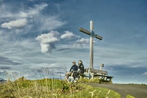 Zwei Personen sitzen auf einer Bank am Gipfelkreuz in Großarl und genießen die Aussicht bei dramatisch bewölktem Himmel. | © Großarler Bergbahnen