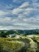 Mountainbiker fährt auf kurvigem Trail mit Blick auf Speicherteich, bewaldete Berghänge und wolkenverhangenen Himmel. | © Großarler Bergbahnen