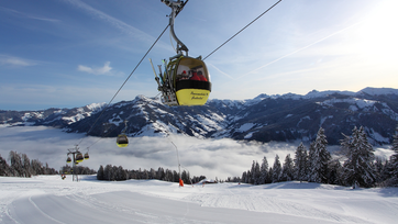 Gelbe Gondeln mit Skifahrern fahren über verschneite Pisten, darunter Wolkenmeer, Berge im Hintergrund, klarer Himmel.