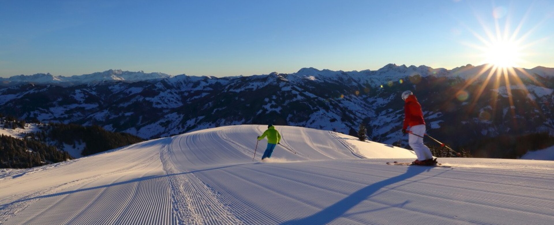 Zwei Skifahrer auf frisch präparierter Piste bei Sonnenaufgang mit Alpenpanorama