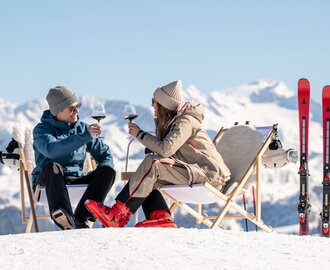 Two skiers sit in deck chairs on snow, toasting red wine, skis beside them with snowy Alpine mountains behind | © Lorenz Masser