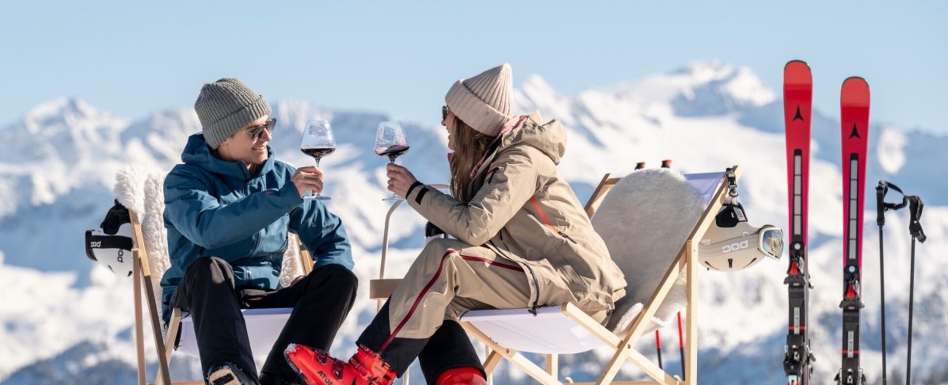 Two skiers sit in deck chairs on snow, toasting red wine, skis beside them with snowy Alpine mountains behind | © Lorenz Masser