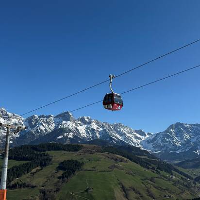 Red Abergbahn gondola glides in summer above green hills and snow-covered Hochkönig range under a clear blue sky. | © Aberg - Hinterthal - Bergbahnen AG