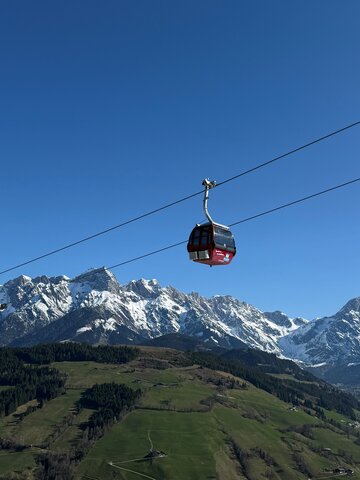 Red Abergbahn gondola glides in summer above green hills and snow-covered Hochkönig range under a clear blue sky. | © Aberg - Hinterthal - Bergbahnen AG