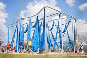 Metal frame with blue fabric loops that can be used as a swing and children playing with it | © Manu Lochner