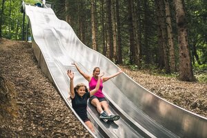Girl and woman laughing with raised arms while sliding down the twin slide at the forest slide park Natrun.