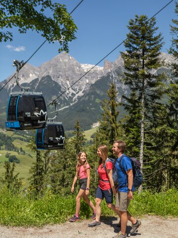 Familie beim Wandern auf Waldweg mit Gondeln im Hintergrund und Bergpanorama | © Roland Haschka
