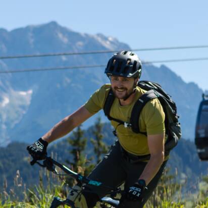 Ein Mountainbiker fährt einen geschwungenen Trail entlang, im Hintergrund sind eine Gondelbahn und hohe Berge sichtbar. | © Roland Haschka