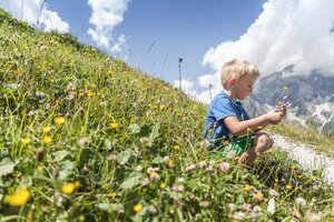 Little boy sits in an alpine meadow picking colorful flowers with mountain scenery and a blue summer sky in the background. | © Felsch Fotodesign