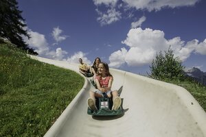 Girl rides the Biberg summer toboggan with two adults behind her, green slope and blue sky in a scenic alpine landscape. | © Hochkönig / Roland Haschka
