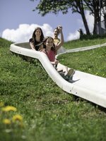 Smiling girl rides the summer toboggan run down a grassy slope with her family, wildflowers visible in the foreground. | © Hochkönig / Roland Haschka