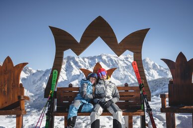 Zwei Personen in Skiausrütung sitzen auf einer Holzbank mit einem Kronen-Rahmen & bestaunen das Panorama | © Christian Schartner