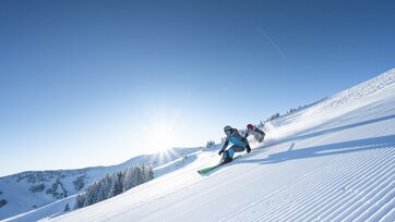 Zwei Skifahrer fahren knapp hintereinander die frisch präparierte Piste hinab und im Hintergrund strahlt die Sonne | © Christian Schartner