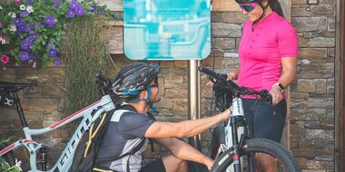 Two cyclists charge their e-bikes at a Hochkönig station surrounded by a sunny alpine setting