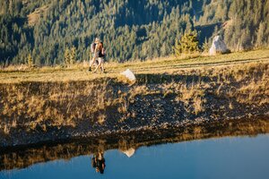 Two hikers walk along the edge of a small pond near Bürglalmbahn in Dienten across a grassy slope. | © Hochkönig Bergbahnen