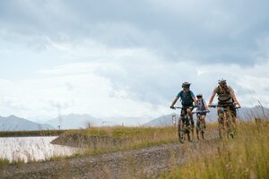 Three mountain bikers ride on a gravel path by a reservoir under cloudy skies near Bürglalmbahn in Dienten | © Hochkönig Bergbahnen