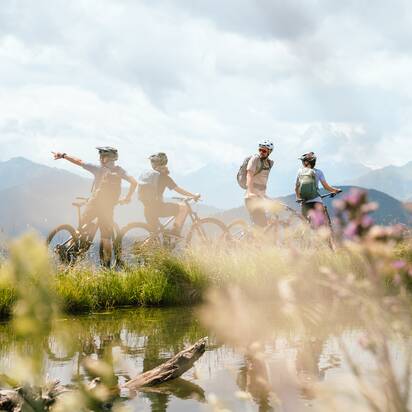Four mountain bikers stand with their bikes by a small pond with mountains behind near Bürglalmbahn in Dienten | © Hochkönig Bergbahnen
