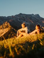Two female hikers sit on a grassy slope in evening light in Dienten with a rocky mountain massif near Gabühelbahn. | © Hochkönig Bergbahnen